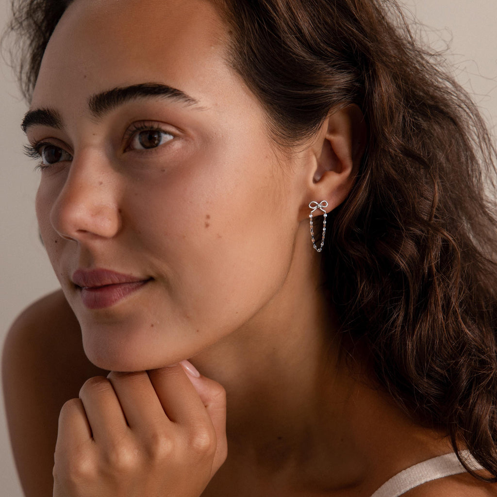 Woman with wavy brown hair wears Bow Chain Studs in Sterling Silver, vintage-inspired bow-shaped earrings, as she rests her chin on her hand.