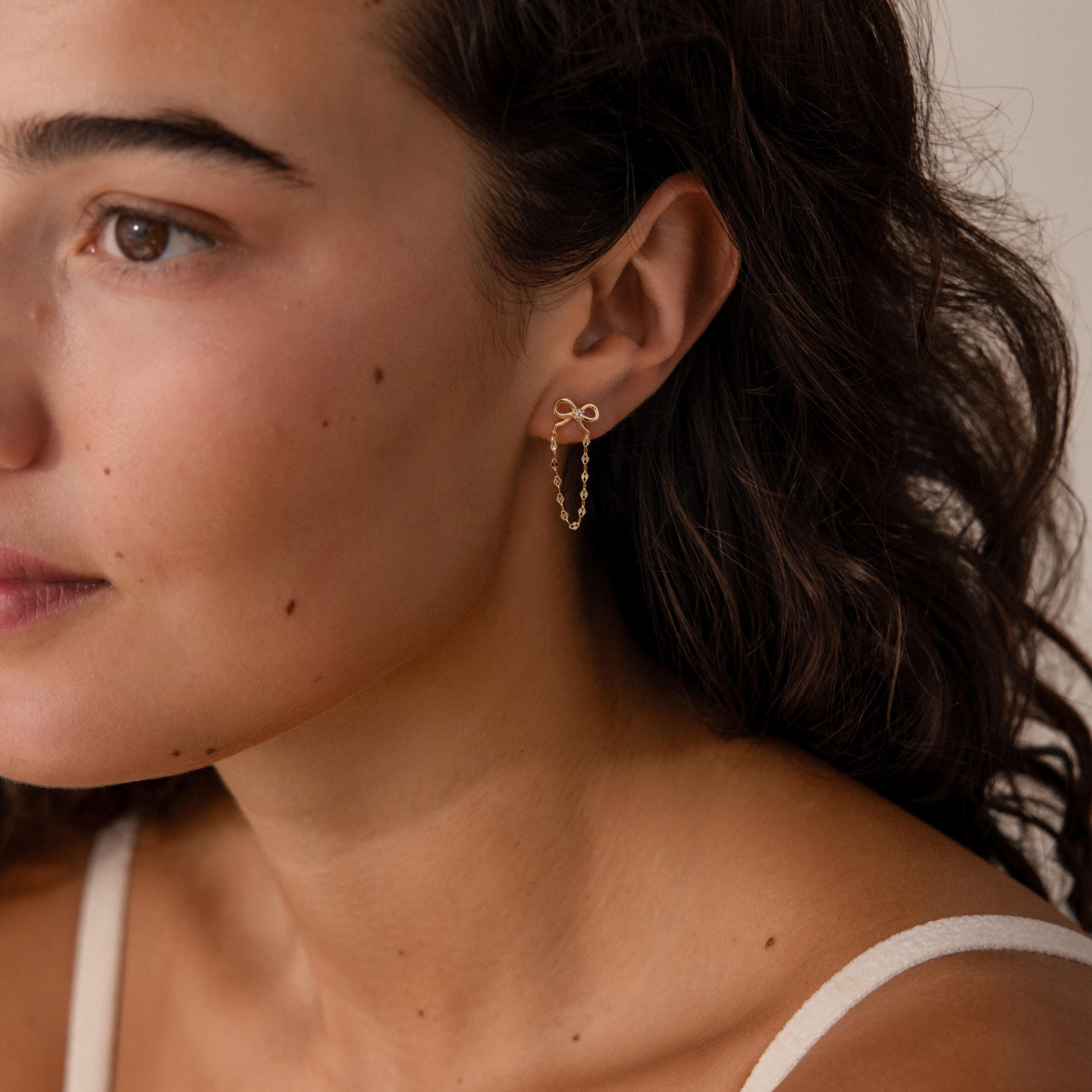 A woman with curly hair wears the Bow Chain Studs in 18K Gold, featuring a delicate gold bow and a small beaded chain, as she looks slightly to the side.