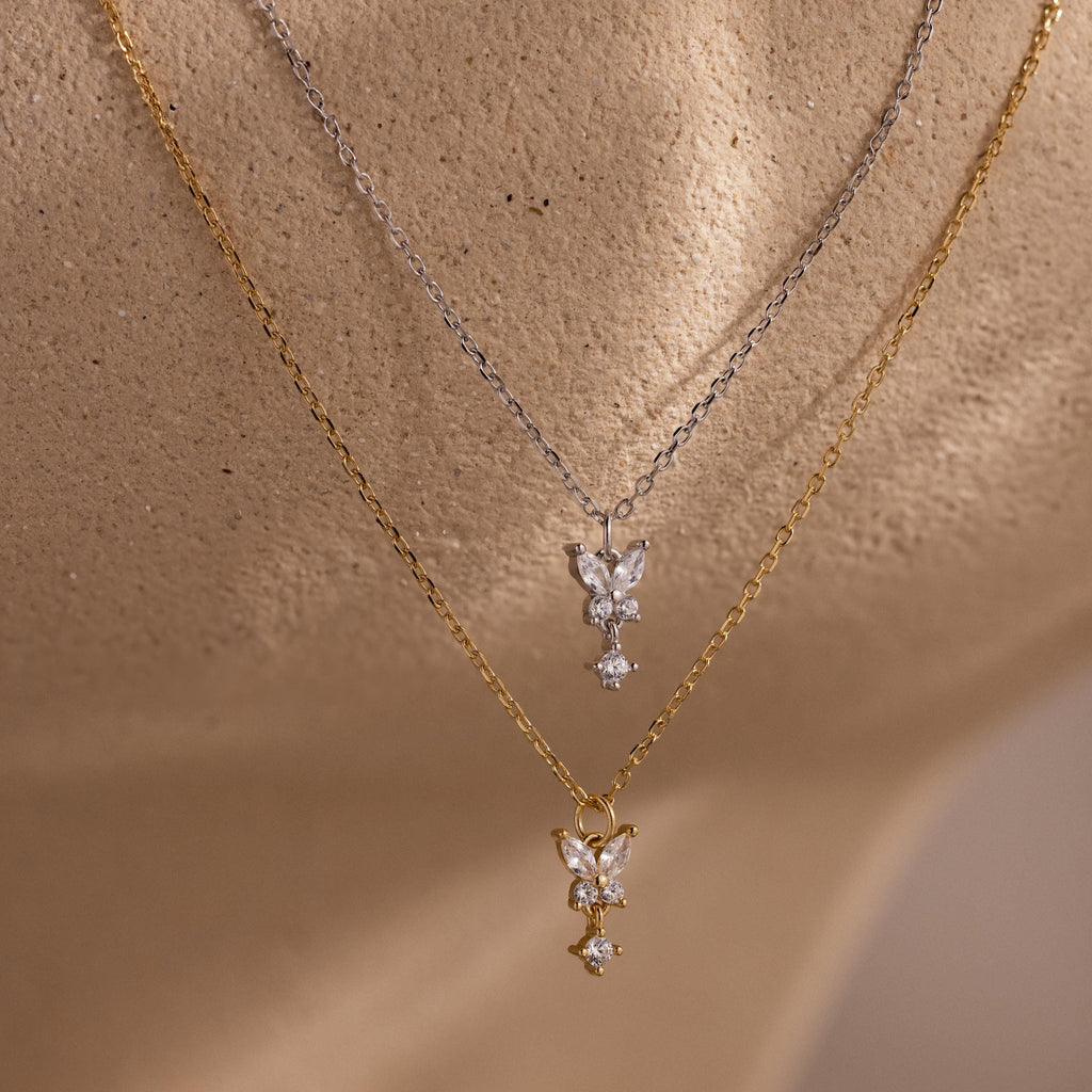 The Butterfly Drop Necklace features two layered chains—one gold and one silver—with butterfly accents, showcasing the elegance of nature-inspired jewelry on a textured surface.