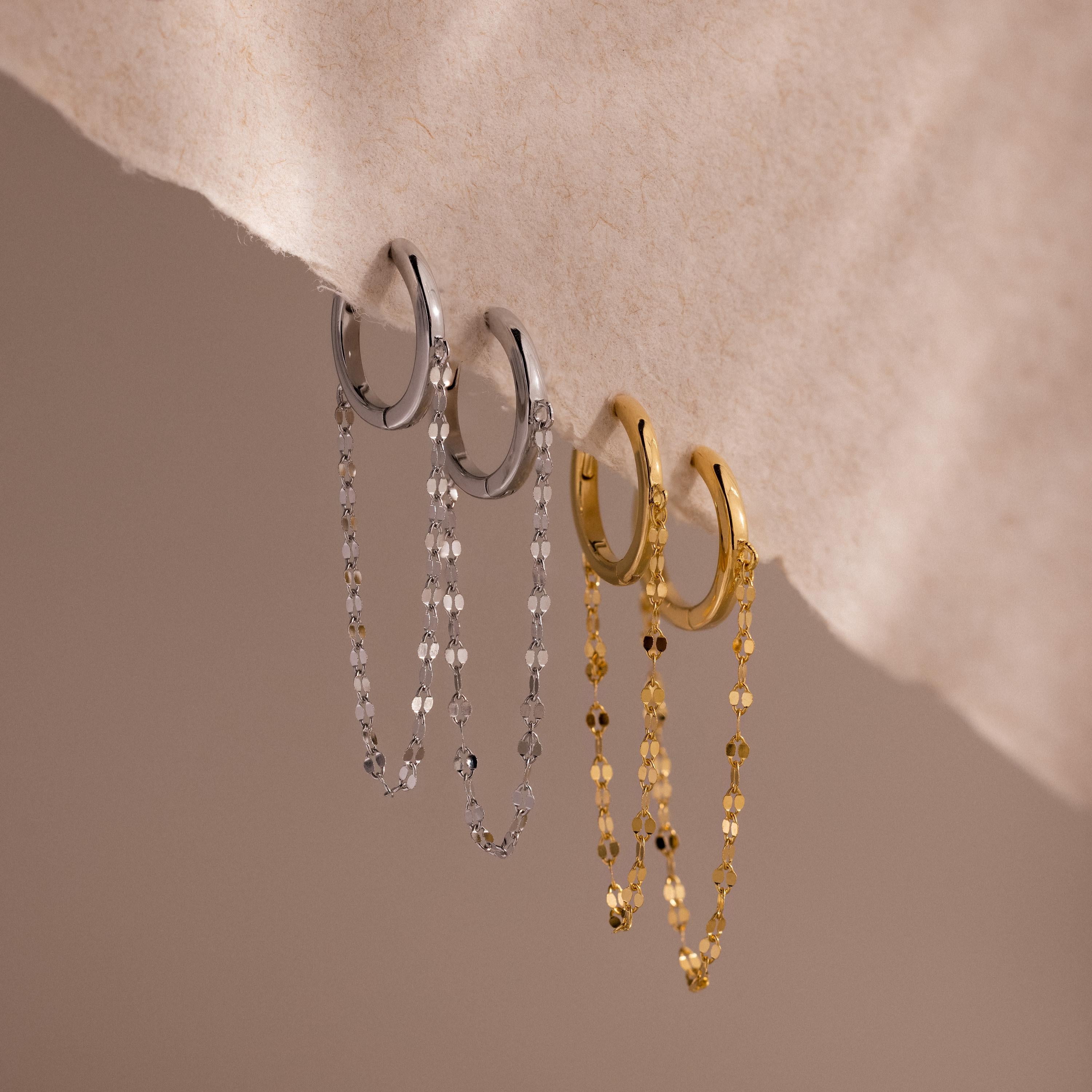 The Mirror Chain Huggies feature two pairs of hoop earrings—one silver, one gold—each with a dangling chain, displayed on soft beige. Perfect minimalist statement jewelry for lovers of bold yet elegant styles.