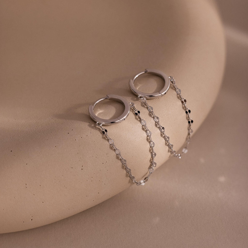 Mirror Chain Huggies in Sterling Silver—statement hoop earrings with delicate chain details, displayed on a smooth beige curved surface.