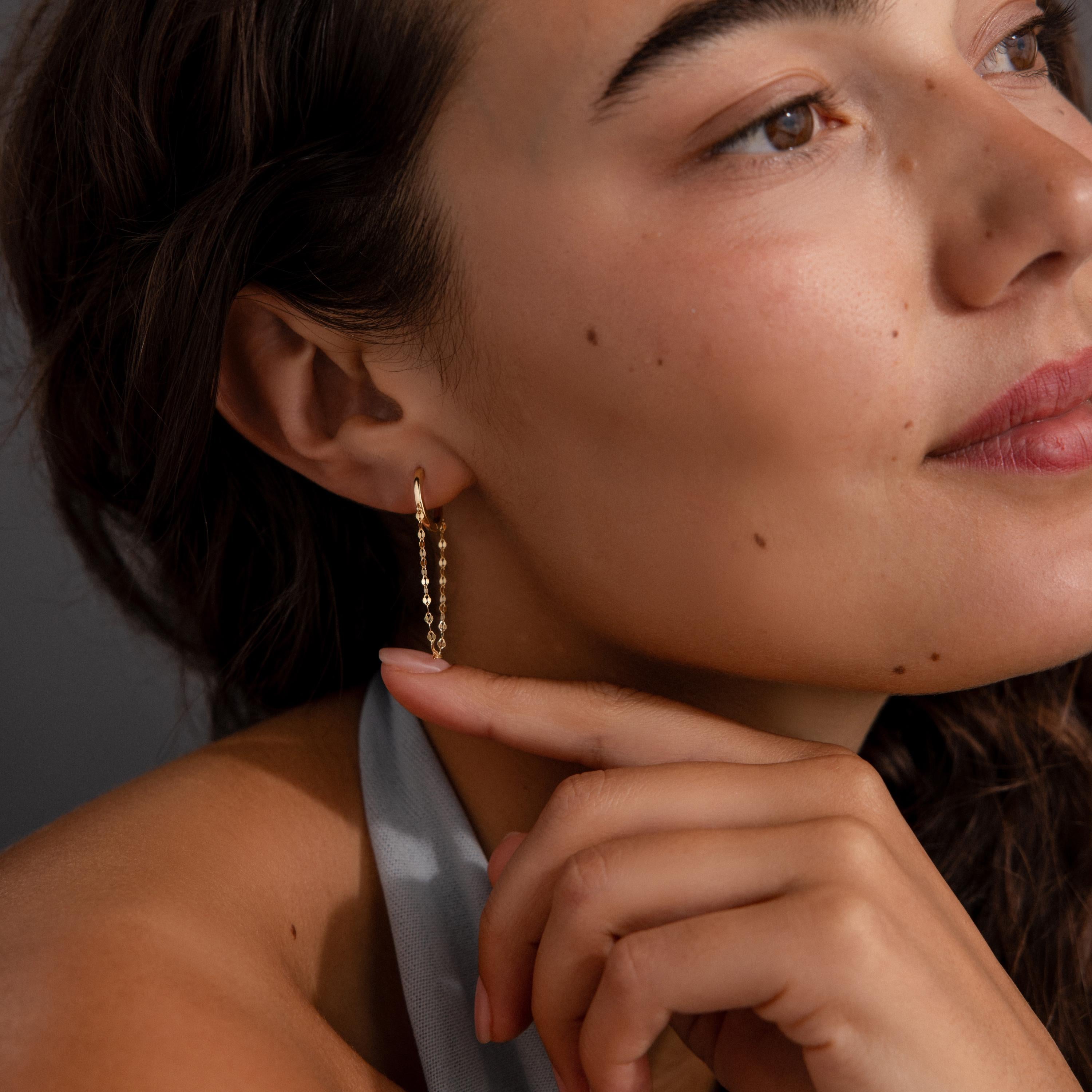 Woman with wavy hair wears Mirror Chain Huggies in 18K Gold, touching her chin and gazing thoughtfully to the side.