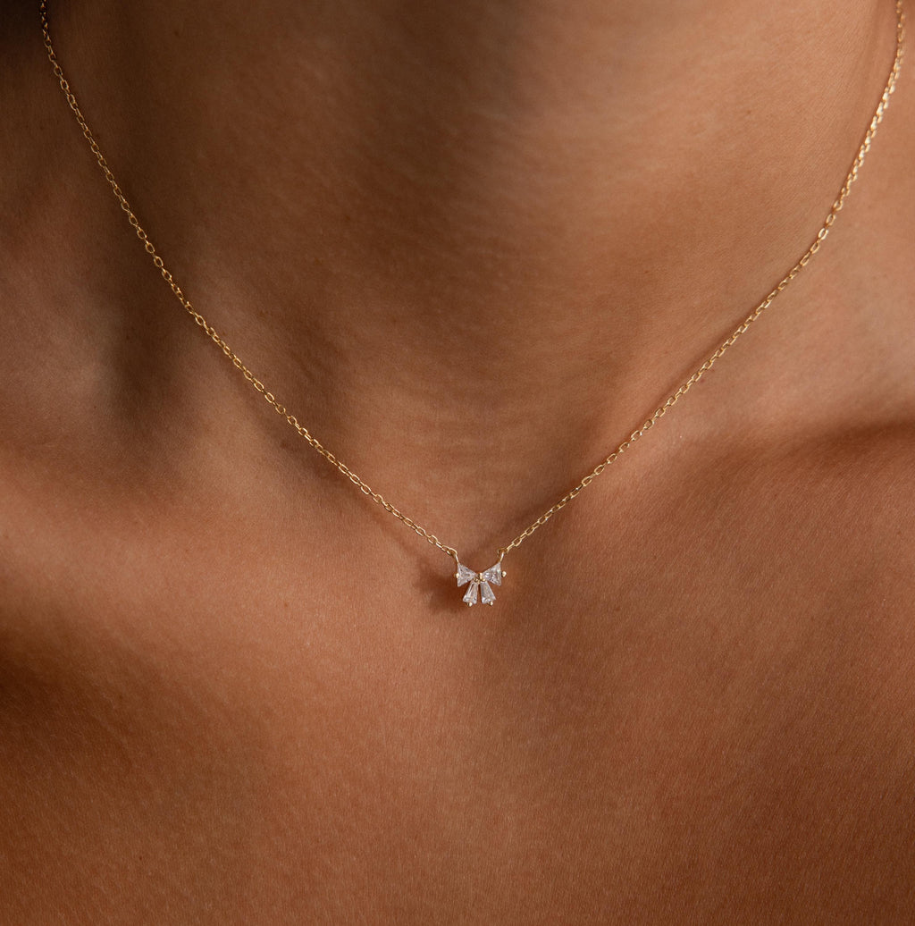 The Diamond Bow Necklace in 18K Gold features a graceful bow design adorned with sparkling stones, elegantly resting on the wearers neck.