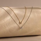 Two elegant Diamond Bow Necklaces, one gold plated and one silver, are displayed on a textured cream surface.