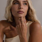 A woman with long blonde hair wears a one-shoulder beige top and gazes upward, her hand adorned with the Zen Diamond Curve Ring resting on her chin.