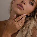 A woman with blonde hair touches her face, wearing the Zen Diamond Curve Ring and other gold rings. She has natural makeup and a neutral expression.