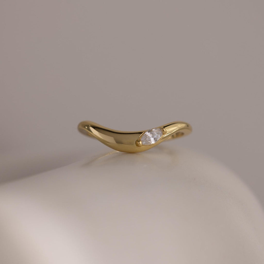The Zen Diamond Curve Ring features a single round marquise diamond accent in a subtle wave design, crafted in gold and displayed on a smooth, off-white surface.