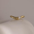 The Zen Diamond Curve Ring features a single round marquise diamond accent in a subtle wave design, crafted in gold and displayed on a smooth, off-white surface.