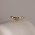 The Zen Diamond Curve Ring features a single round marquise diamond accent in a subtle wave design, crafted in gold and displayed on a smooth, off-white surface.