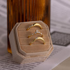 The Zen Diamond Curve Ring with a marquise diamond accent and two minimalist gold curve rings are displayed on a beige velvet holder in front of a brown glass bottle atop printed papers.