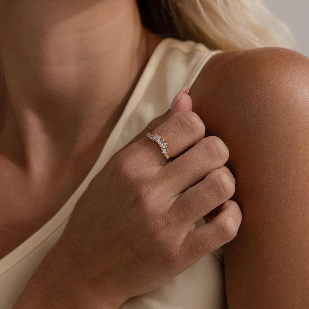 A person with light skin touches their collarbone while wearing the Diamond Curve Cluster Ring, featuring several small clear gemstones in different shapes on their ring finger.