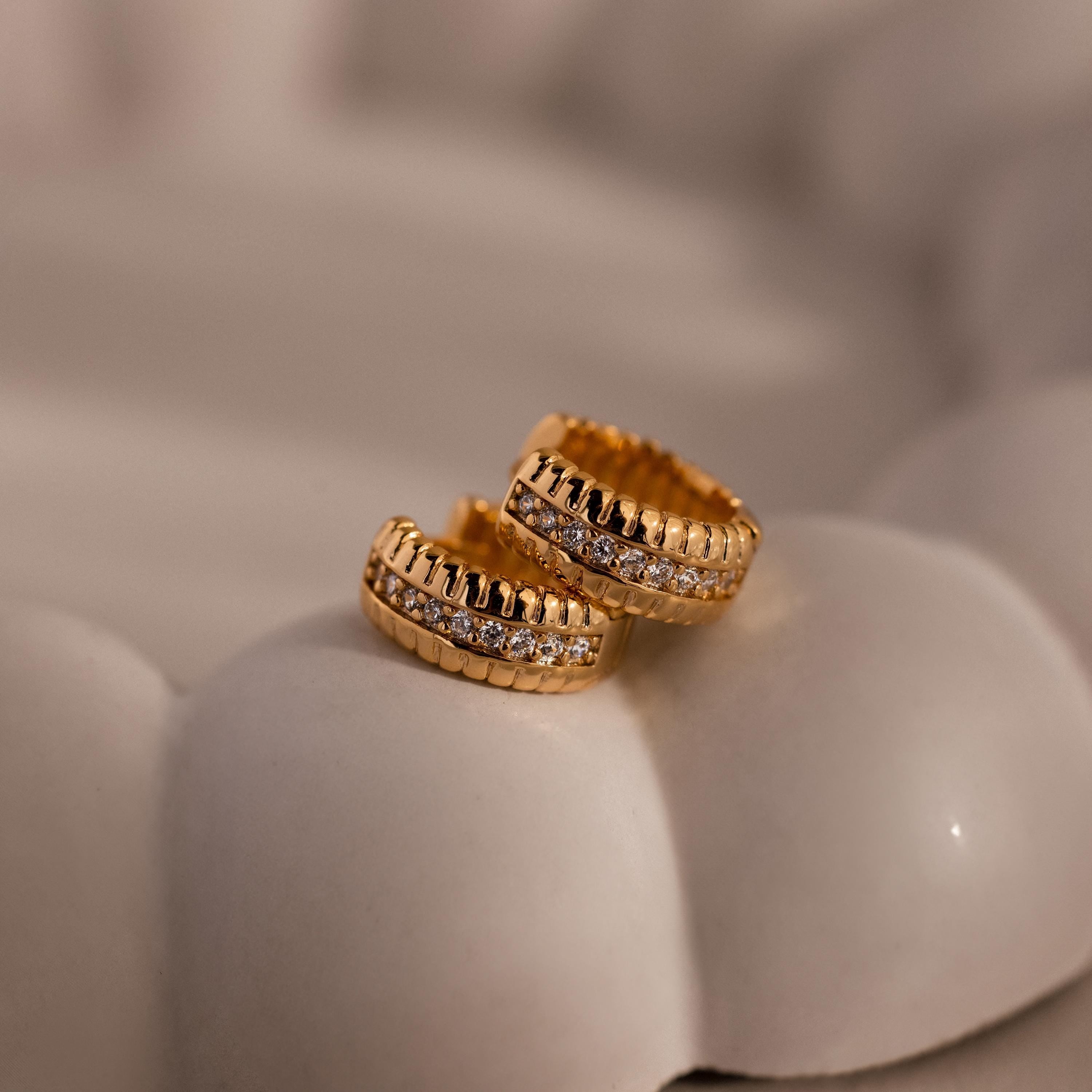 Two Ribbed Pave Huggies in 18K Gold, featuring embedded clear stones, rest on a soft white cushion—ideal as elegant cubic zirconia earrings for any occasion.