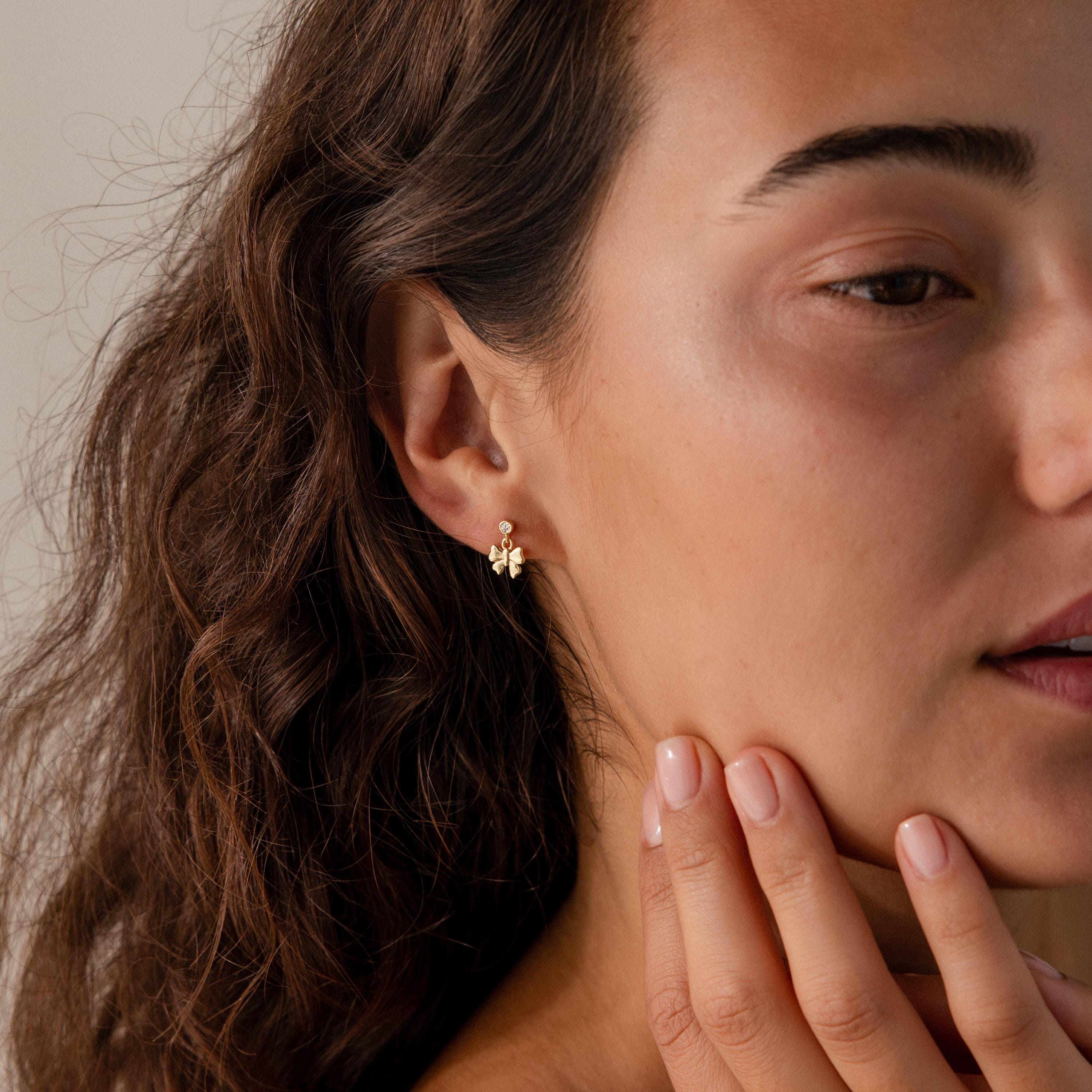 Woman with wavy brown hair wears delicate Bow Drop Studs in 18K Gold, gently touching her face with her hand.