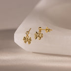 The Bow Drop Studs in 18K Gold, perfect for anyone who loves charming jewelry, are beautifully displayed on a smooth, light-colored surface.