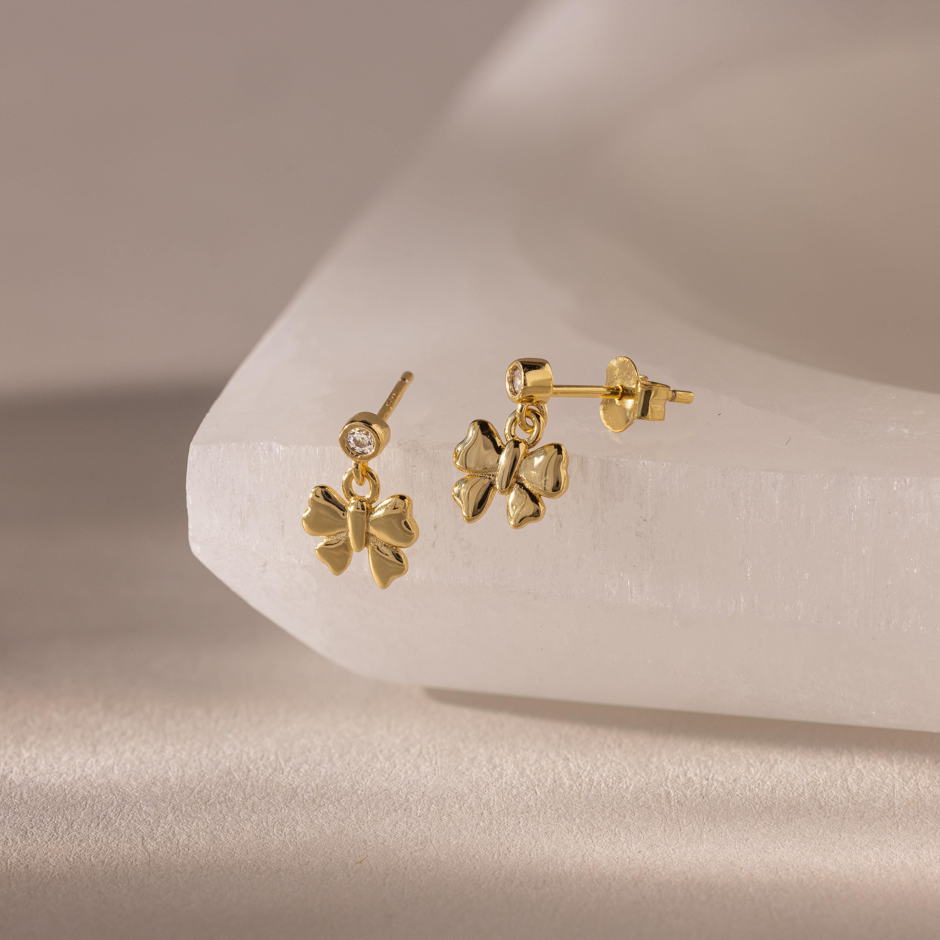 The Bow Drop Studs in 18K Gold, perfect for anyone who loves charming jewelry, are beautifully displayed on a smooth, light-colored surface.