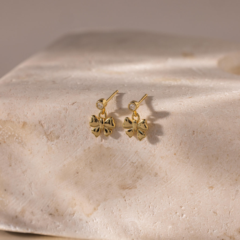 Bow Drop Studs are elegant earrings featuring small gemstones. Beautifully displayed on a textured light stone surface.