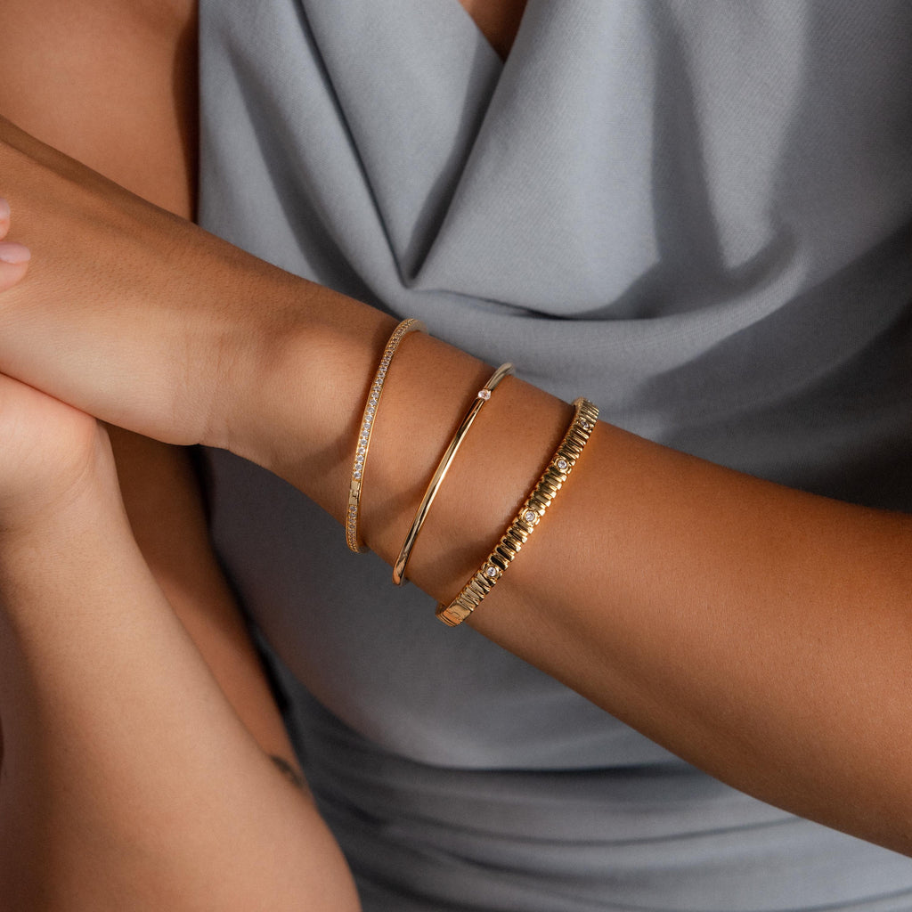 A woman in a light gray sleeveless top rests her arms on a surface, wearing three gold bangles, including the Skinny Diamond Bangle—a minimalist bracelet with sparkling cubic zirconia.