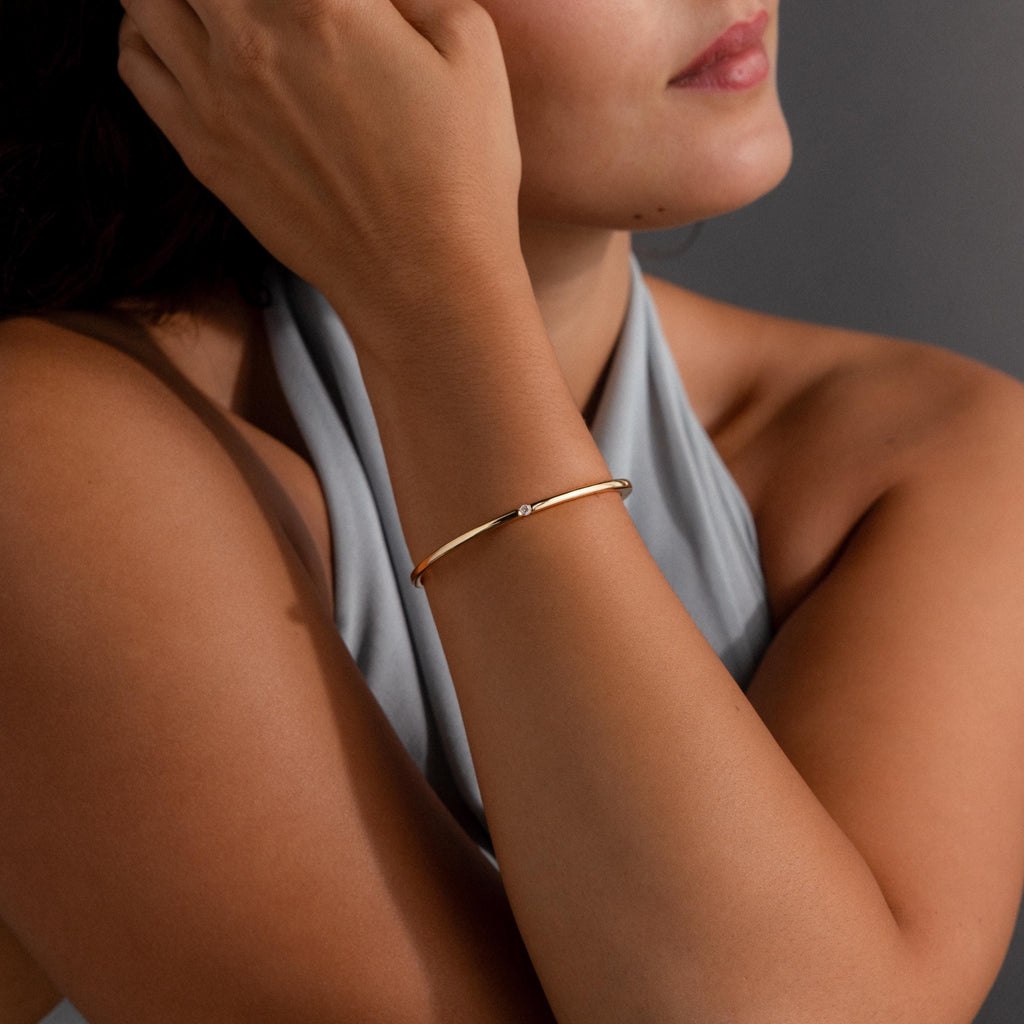 A woman wearing a sleeveless gray top and the Skinny Diamond Bangle—a minimalist gold bracelet featuring a small cubic zirconia gemstone.