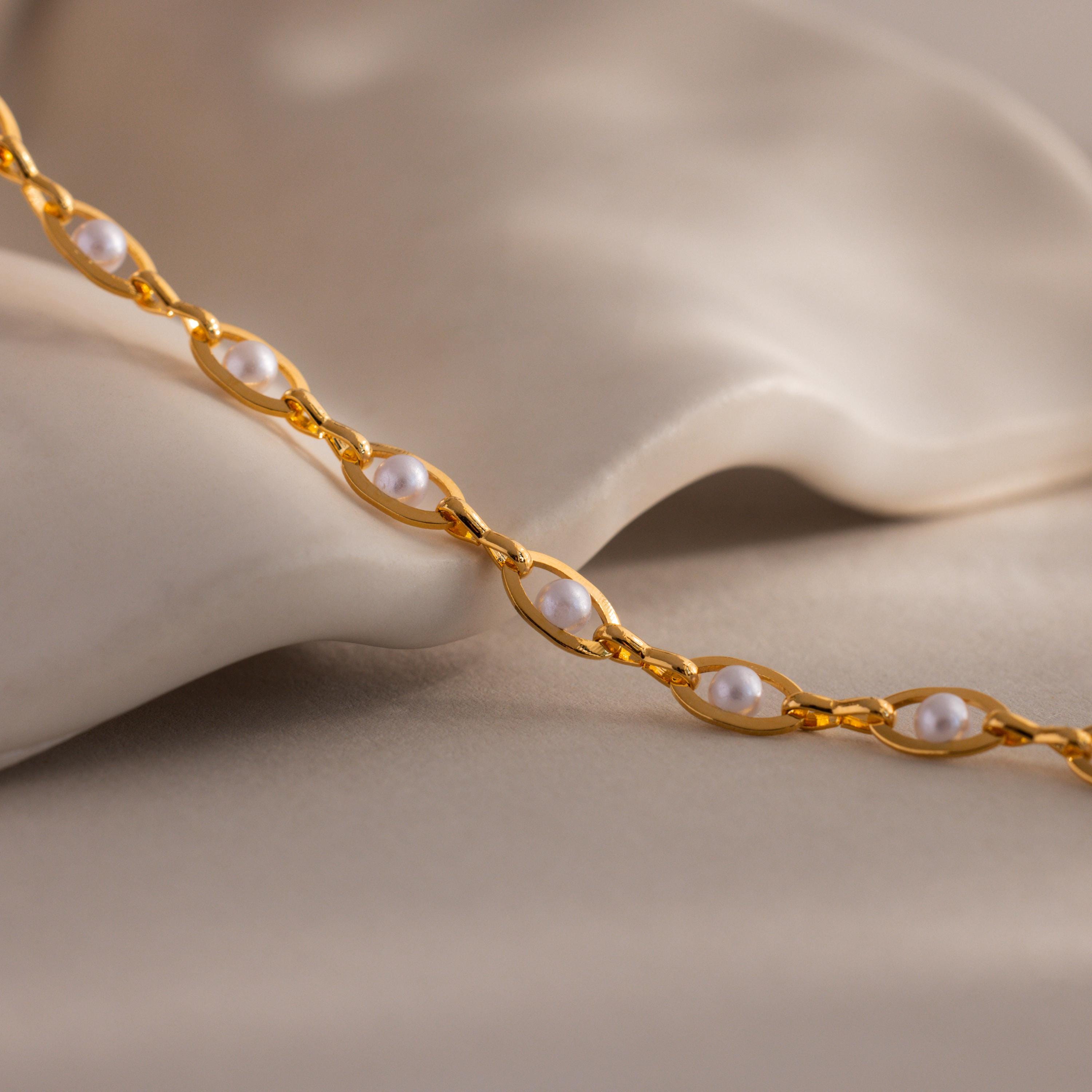 Close-up of a gold chain bracelet with white pearls set in oval links, displayed against a soft neutral fabric background.