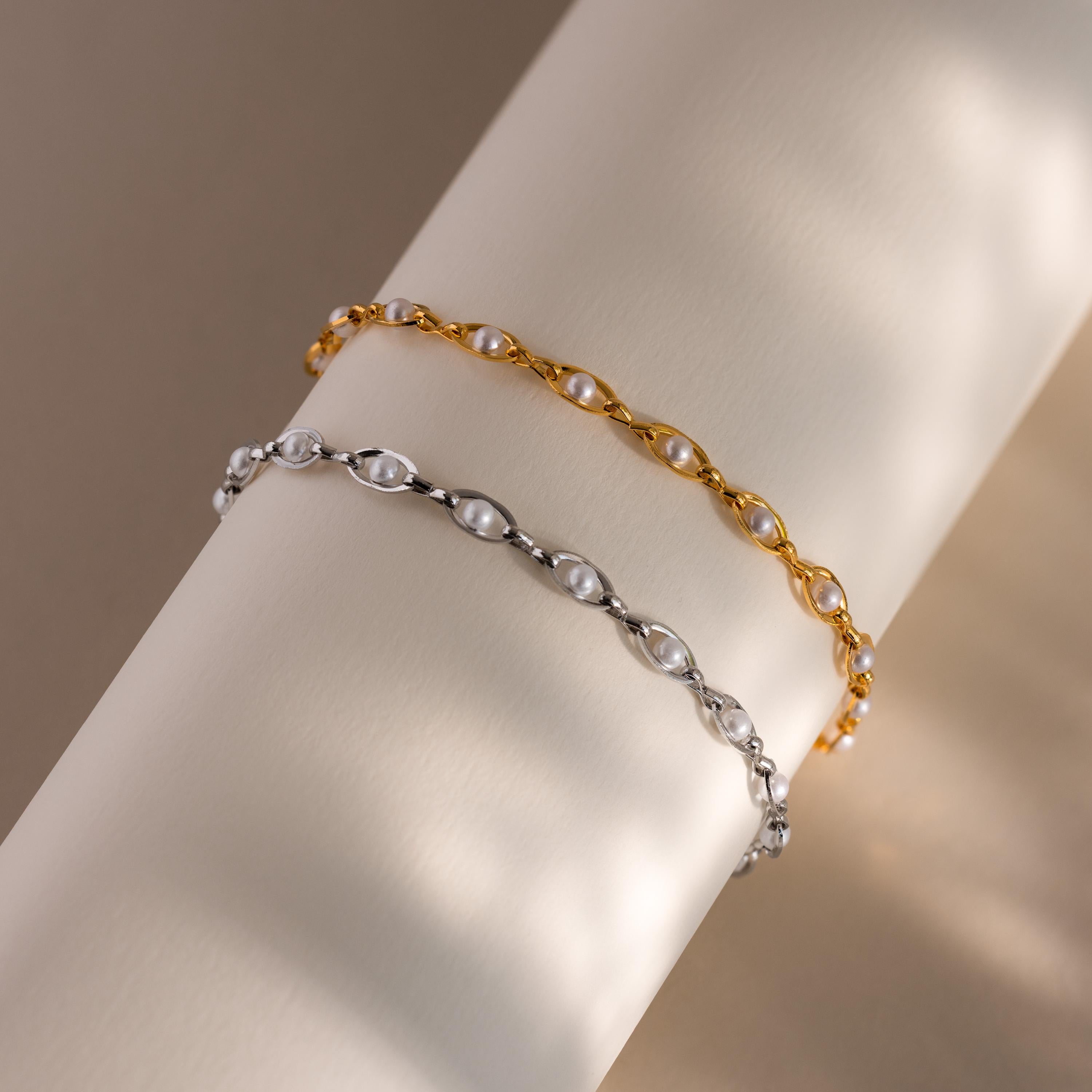 Gold and silver chain bracelets with evenly spaced white pearls, displayed side by side on a neutral cylindrical surface with soft light reflections.