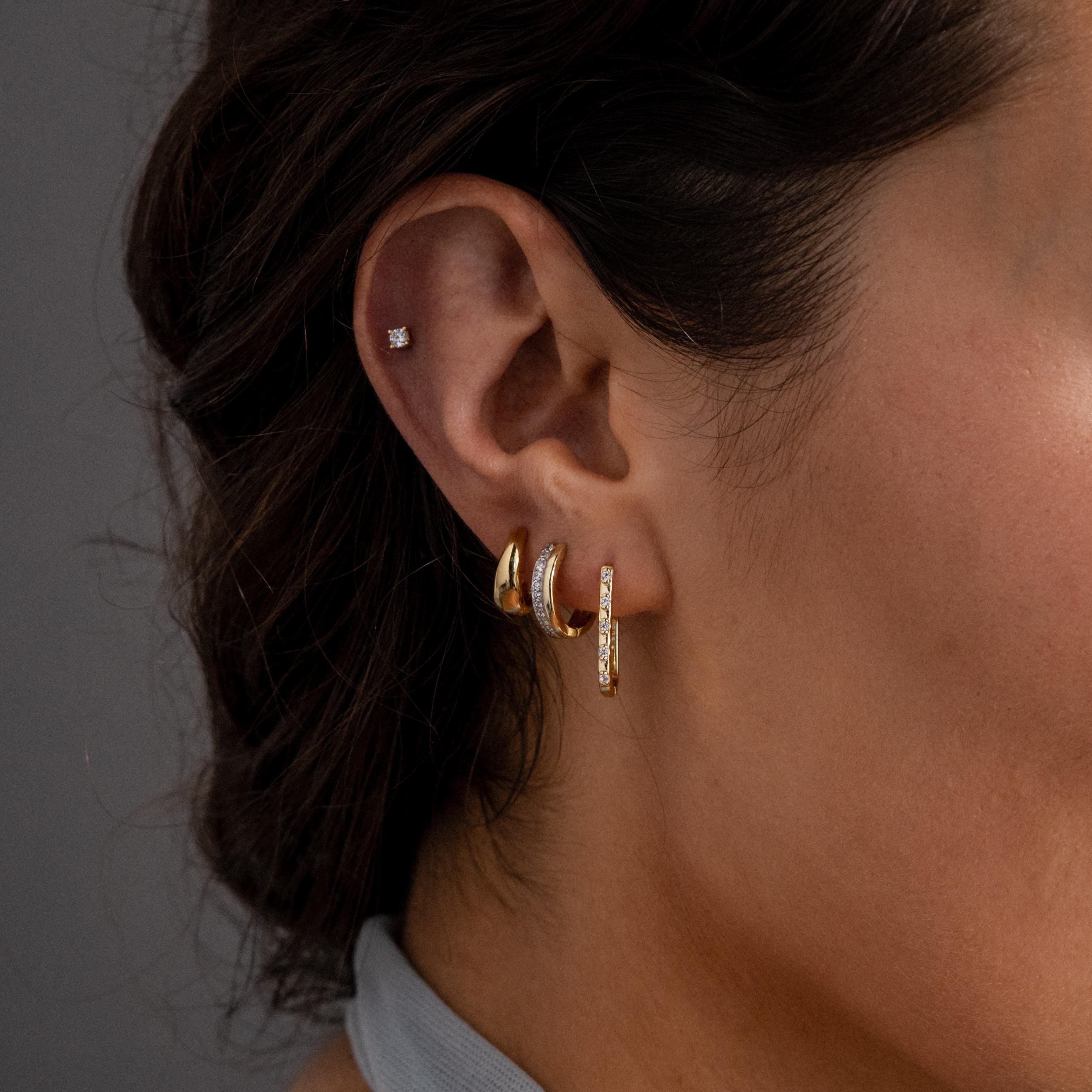 Close-up of a woman’s ear wearing four minimalist earrings, including Dainty Pave Link Earrings in 18K Gold, a small stud, and two elegant gold hoops.