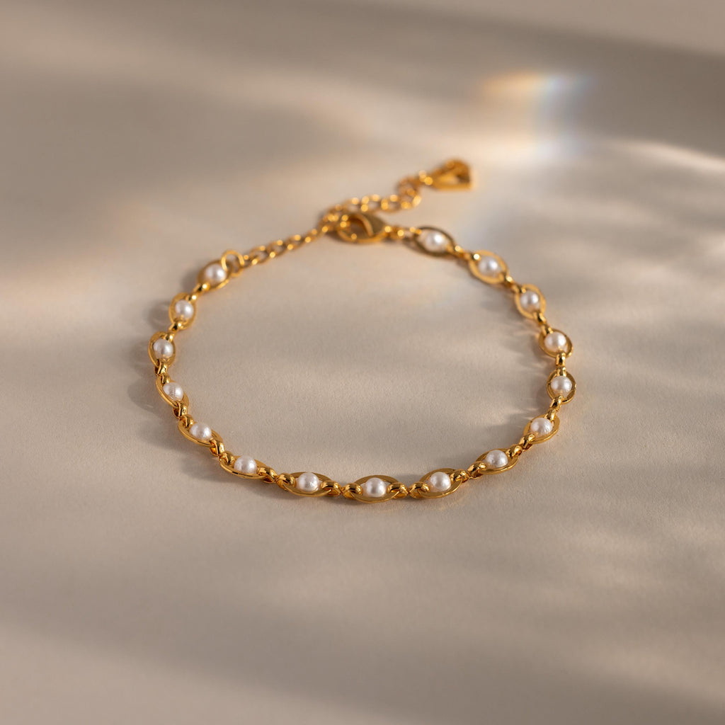 Gold chain bracelet with evenly spaced white pearls, featuring a delicate adjustable clasp and heart-shaped detail, displayed on a neutral surface with soft light reflections.