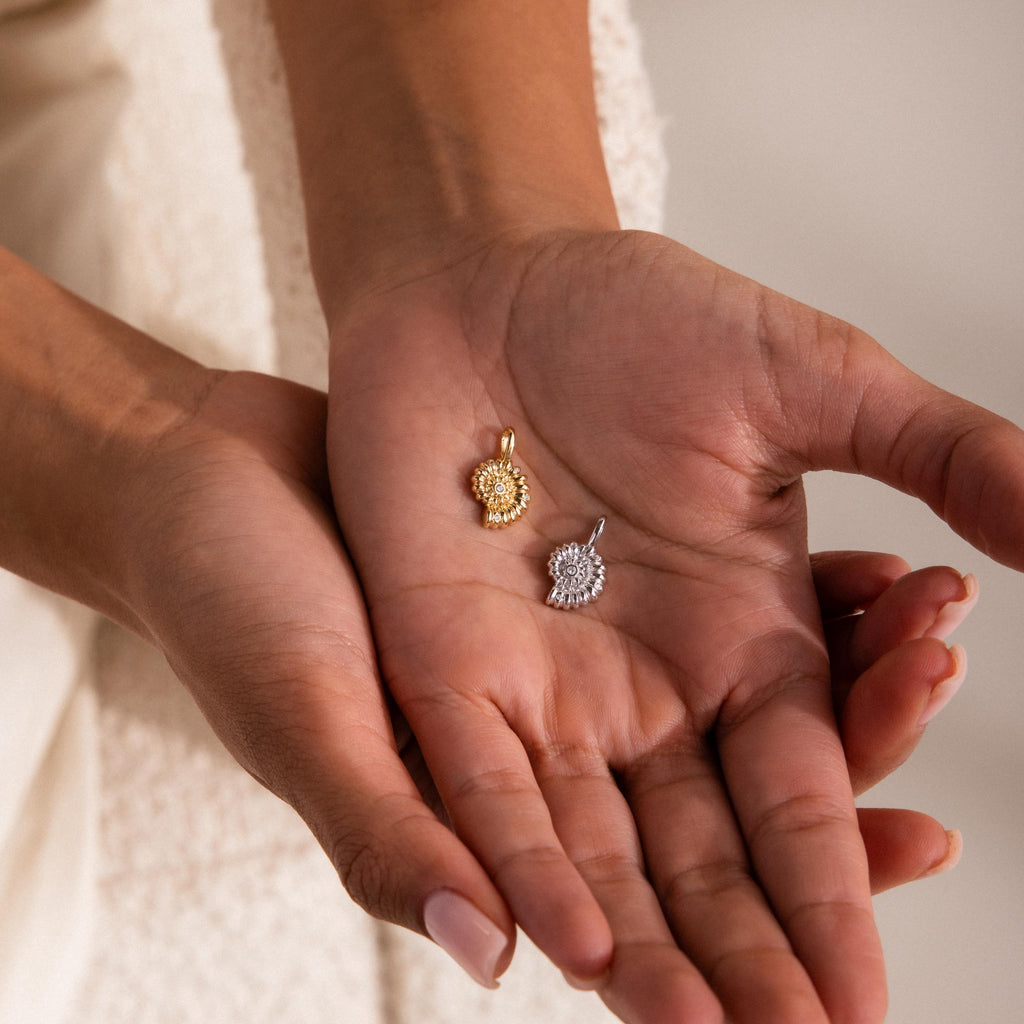 Gold and silver shell-inspired charm pendants with textured circular designs and gemstone accents, displayed in the palm of a hand.