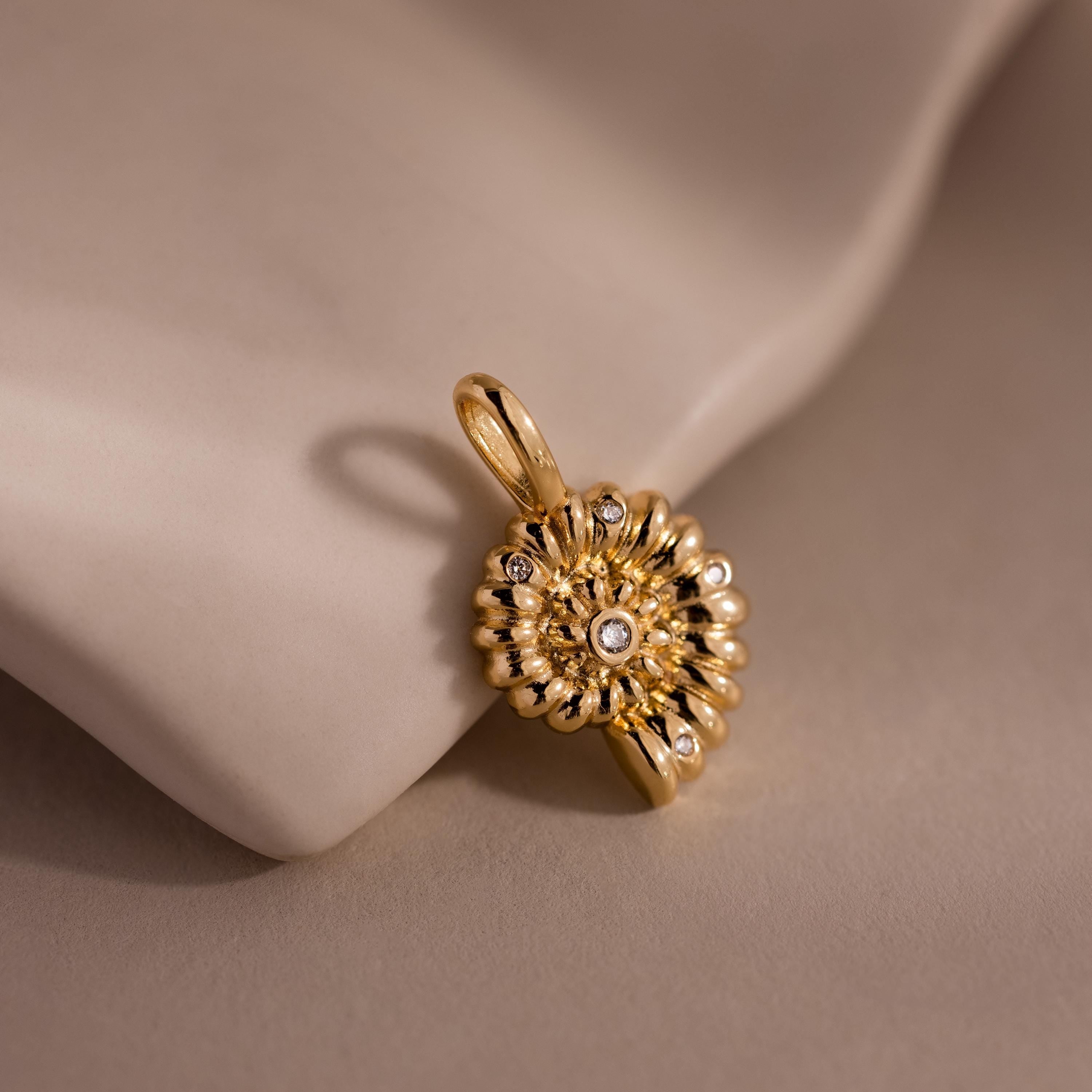 Gold shell inspired charm pendant with a textured circular design, accented by small white gemstones, displayed on a soft neutral fabric background.