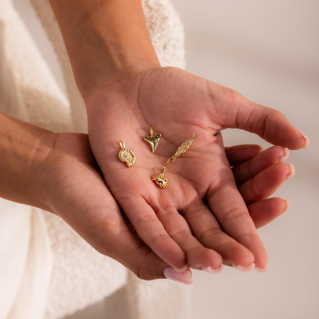 Gold ocean-inspired charm pendants including a shell, shark tooth, fish, and crab design, displayed in the palm of a hand.
