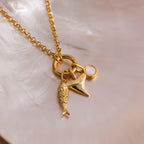 Gold chain necklace with three charms: a detailed fish, a shark tooth pendant, and a round white stone, displayed on an iridescent seashell background.