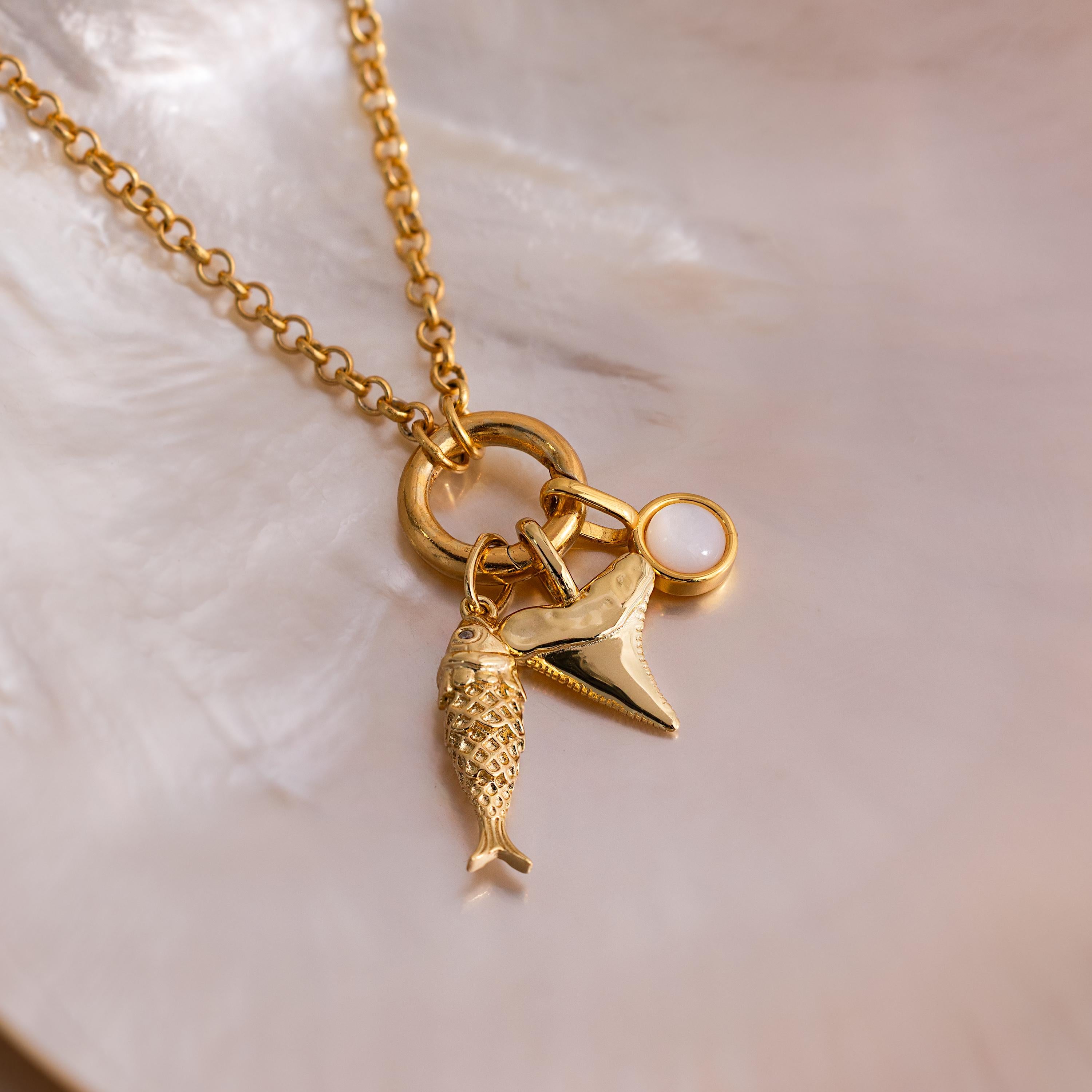 Gold chain necklace with three charms: a detailed fish, a shark tooth pendant, and a round white stone, displayed on an iridescent seashell background.