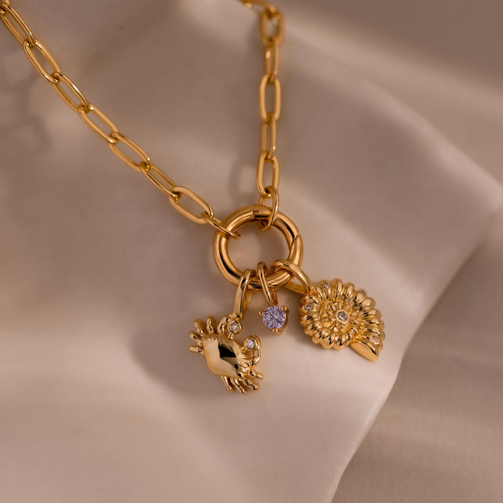 Gold chain necklace with three charms: a crab-shaped charm with gemstone eyes, a small purple gemstone, and a shell-inspired pendant with crystal accents, displayed on a soft neutral fabric background.