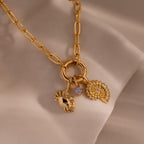 Gold chain necklace with three charms: a crab-shaped charm with gemstone eyes, a small purple gemstone, and a shell-inspired pendant with crystal accents, displayed on a soft neutral fabric background.