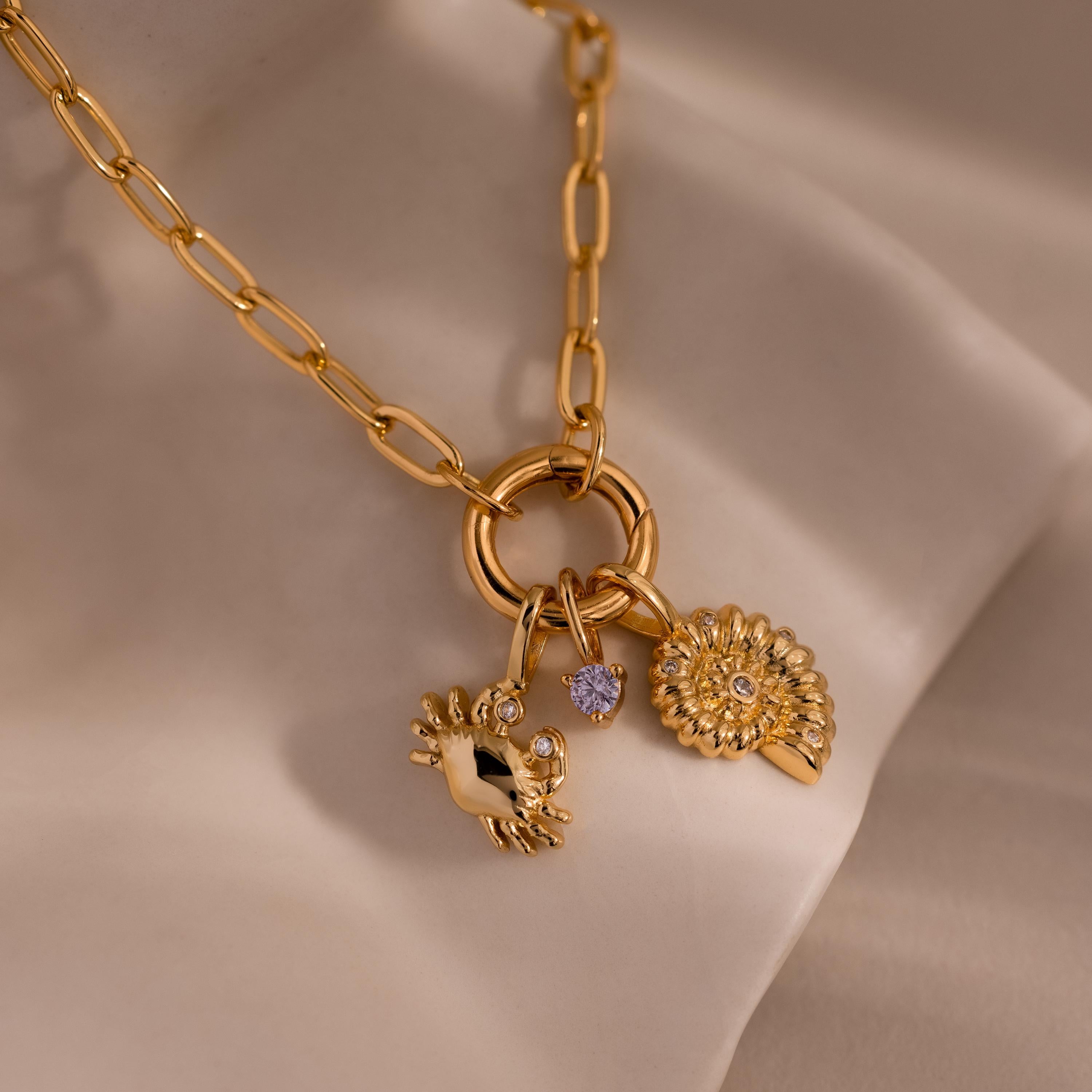 Gold chain necklace with three charms: a crab-shaped charm with gemstone eyes, a small purple gemstone, and a shell-inspired pendant with crystal accents, displayed on a soft neutral fabric background.