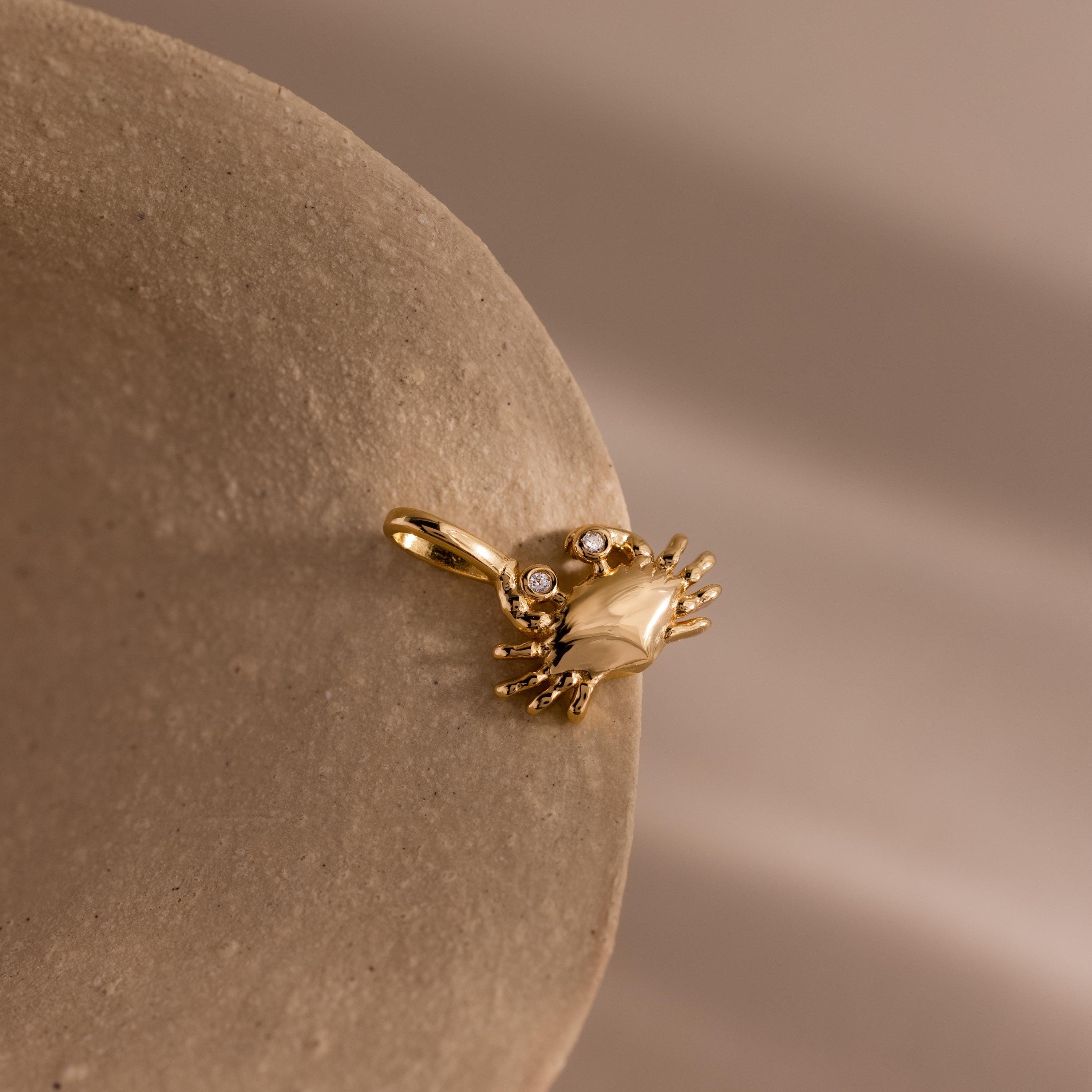Gold crab shaped charm pendant with gemstone accented eyes, displayed on a smooth stone surface against a neutral background.