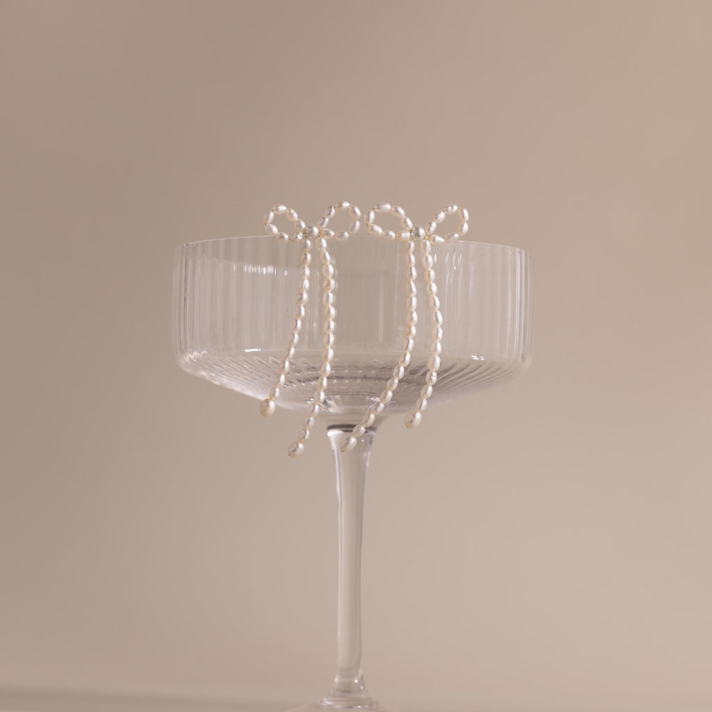 The Pearl Bow Earrings, shaped like delicate bows, hang on the rim of a clear ribbed glass coupe set against a beige background—an elegant jewelry statement.