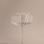 The Pearl Bow Earrings, shaped like delicate bows, hang on the rim of a clear ribbed glass coupe set against a beige background—an elegant jewelry statement.