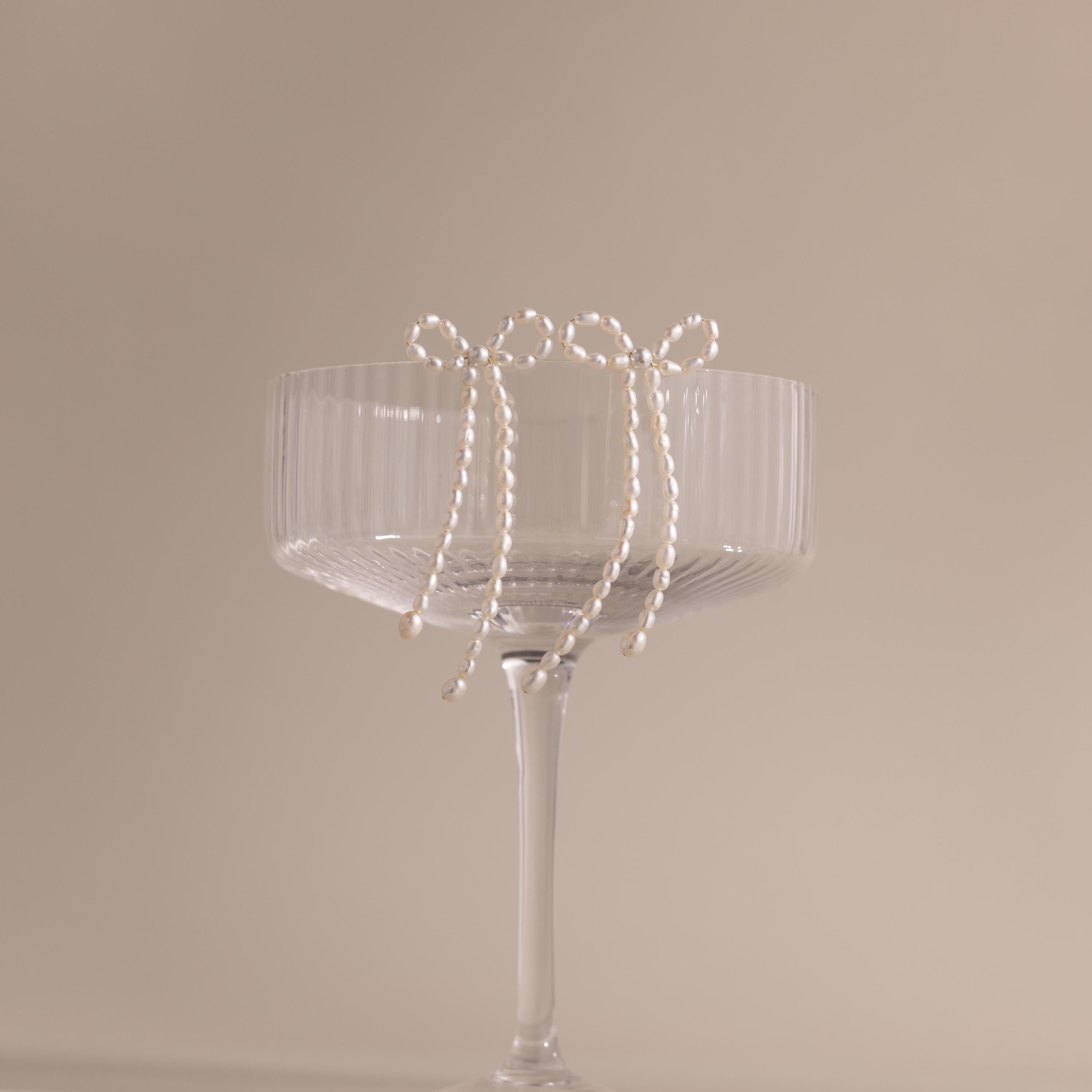 The Pearl Bow Earrings, shaped like delicate bows, hang on the rim of a clear ribbed glass coupe set against a beige background—an elegant jewelry statement.