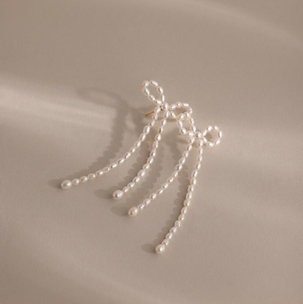 Pearl Bow Earrings feature a chic bow design with three delicate strands, making a stylish statement on a beige background.
