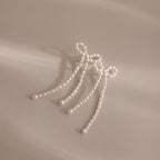 Pearl Bow Earrings feature a chic bow design with three delicate strands, making a stylish statement on a beige background.