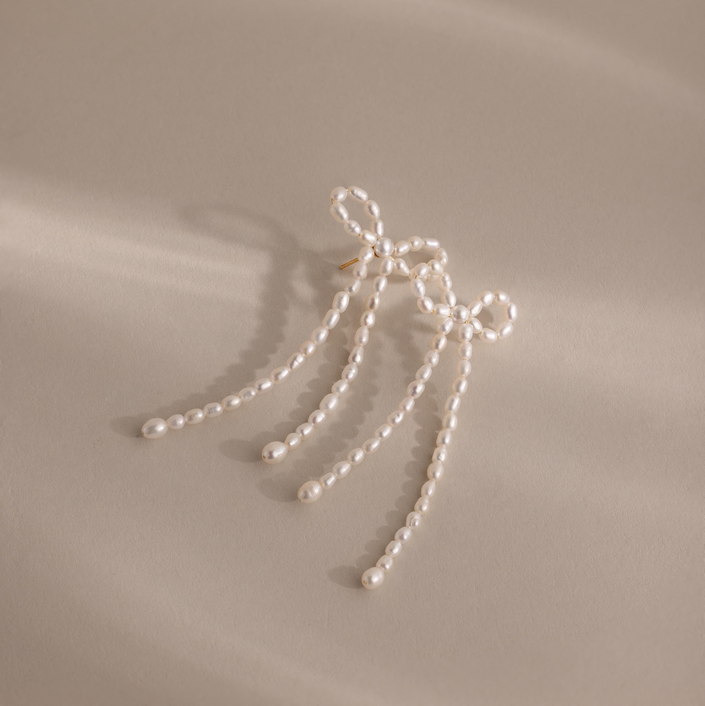 Pearl Bow Earrings feature a chic bow design with three delicate strands, making a stylish statement on a beige background.