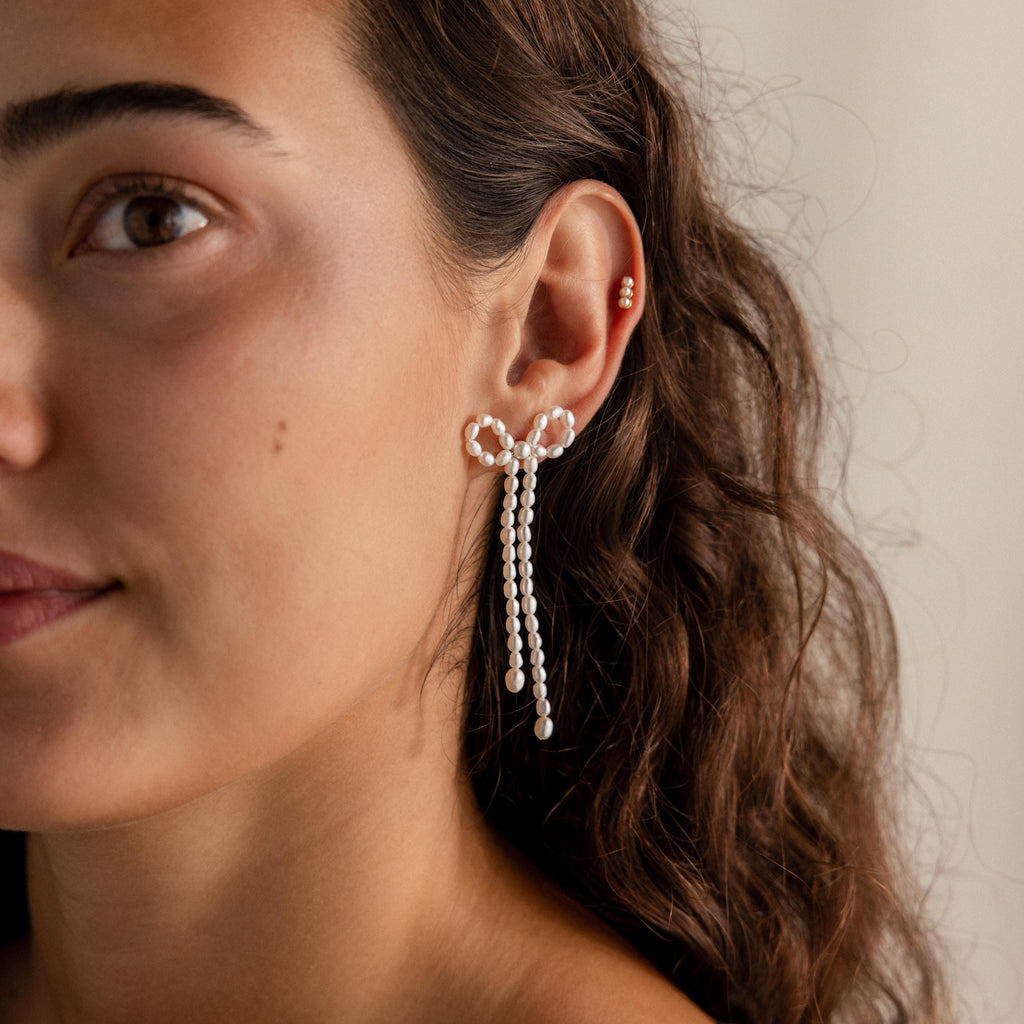 Woman with wavy brown hair wearing elegant Pearl Bow Earrings and a small gold stud on her ear.