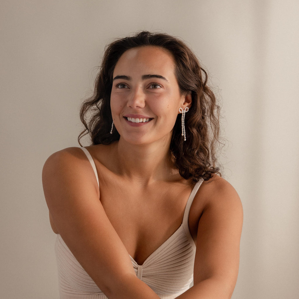 A woman with wavy brown hair smiles in a beige top, showcasing Pearl Bow Earrings against a neutral background.