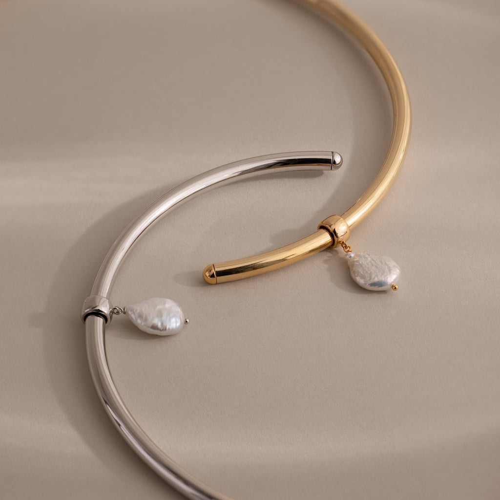The Pearl Cuff Choker Necklace comes in silver and gold tones, each featuring a curved metal band with a dangling baroque pearl charm for an elegant touch.