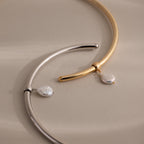 The Pearl Cuff Choker Necklace comes in silver and gold tones, each featuring a curved metal band with a dangling baroque pearl charm for an elegant touch.
