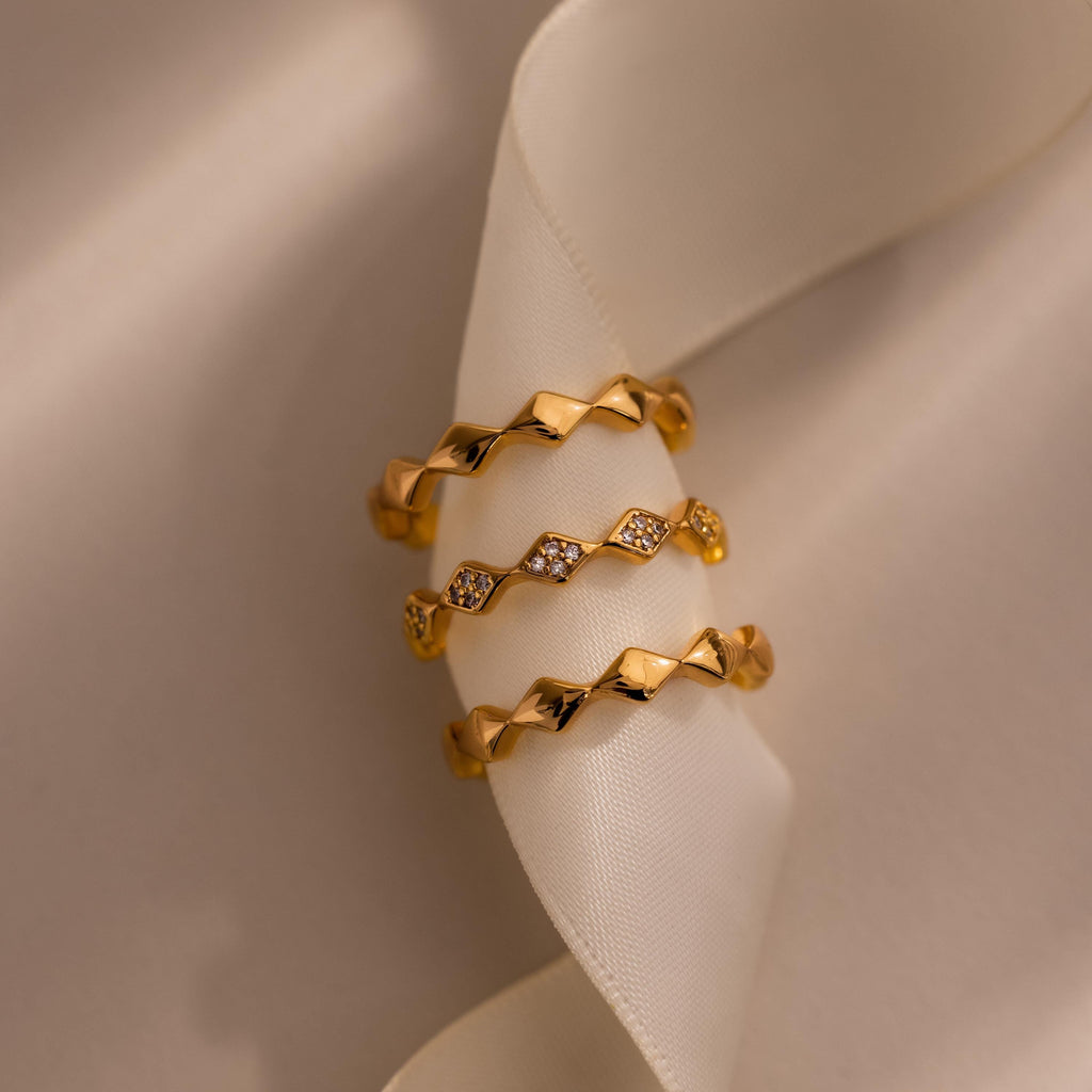 Set of three eternity curve rings in gold, featuring quilted wave design with one band accented by sparkling diamonds, displayed on soft ivory fabric.
