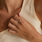 Gold heart curve cluster diamond ring with heart shaped center stone and round diamonds worn on a woman’s hand against a beige top.