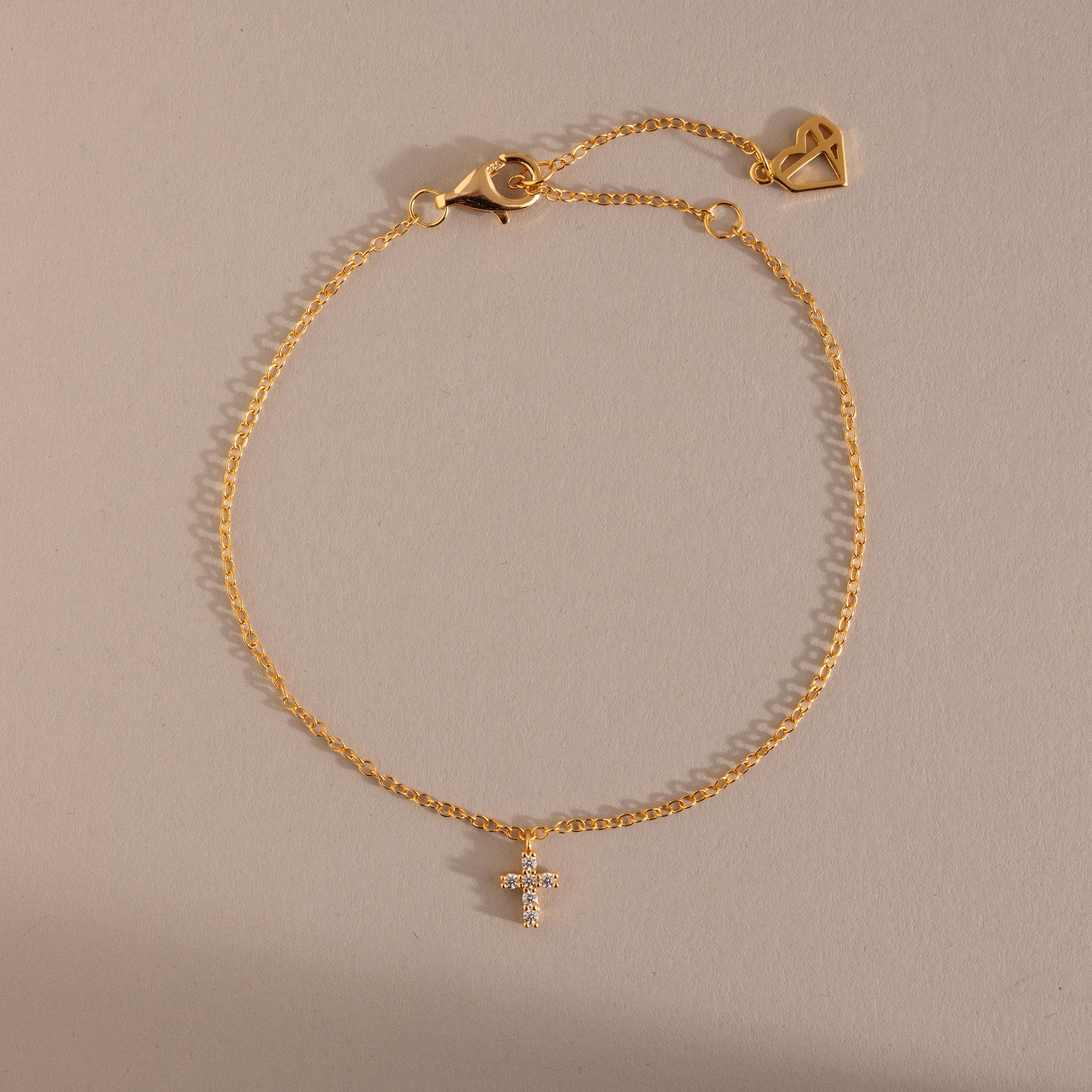 Gold chain bracelet featuring a small crystal cross charm and adjustable clasp with heart detail, a delicate and meaningful jewelry piece perfect for layering or everyday wear.