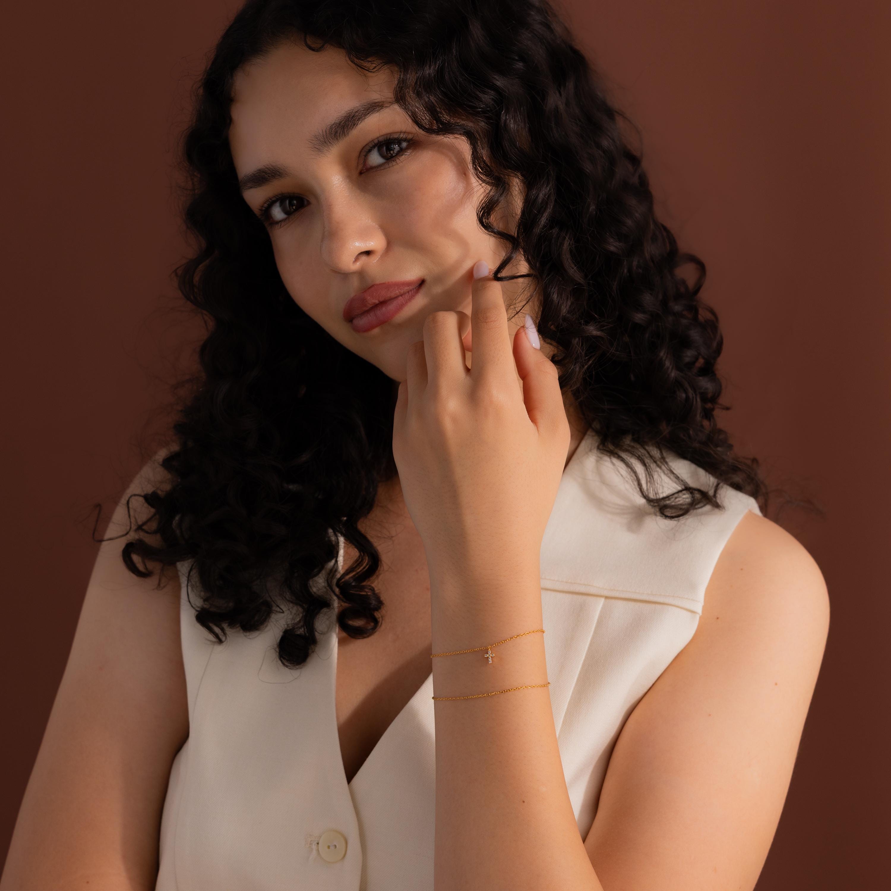 Woman wearing a gold chain bracelet with a delicate crystal cross charm, styled with a sleeveless cream blouse against a warm brown background for an elegant look.