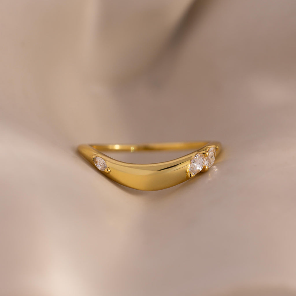 Close up of a gold curved band ring with three marquise-cut white gemstones, displayed on soft beige fabric for a minimal and elegant jewelry presentation.